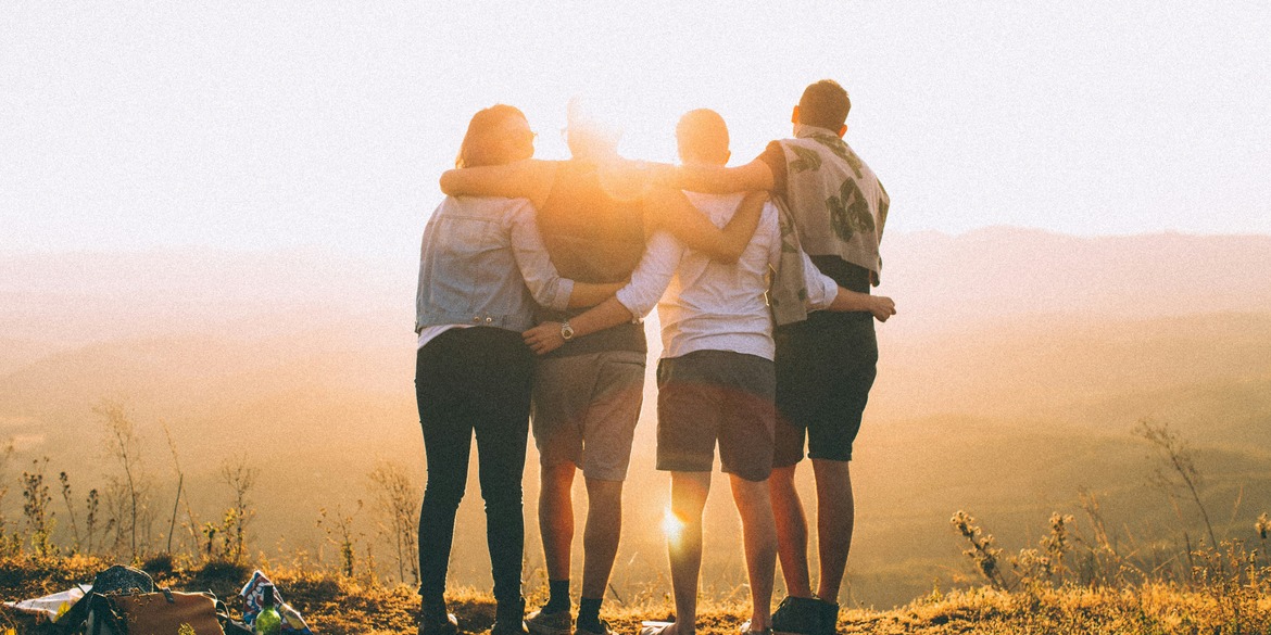4 individuals stand in a close-knit circle, their backs turned towards the camera as they embrace one another, captivated by a mountain panorama