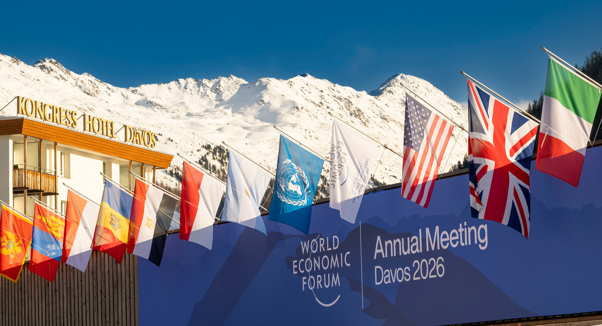 Photo of entrance to World Economic Forum, Davos, showing several European flags with a snowy mountain backdrop.