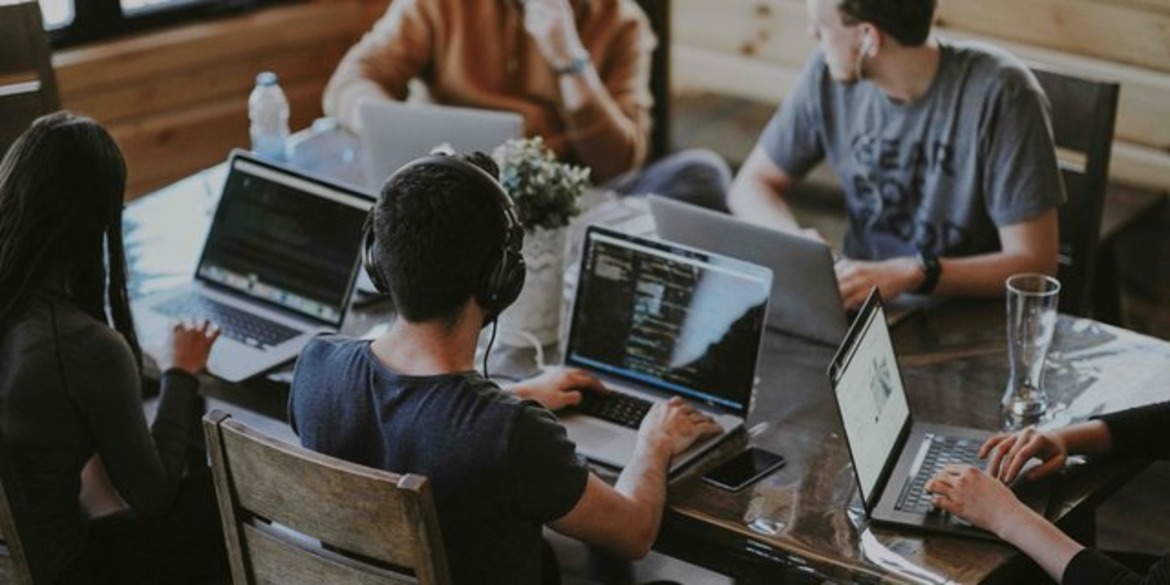 Photo of young adults sitting around a table on their laptops - some with headphones