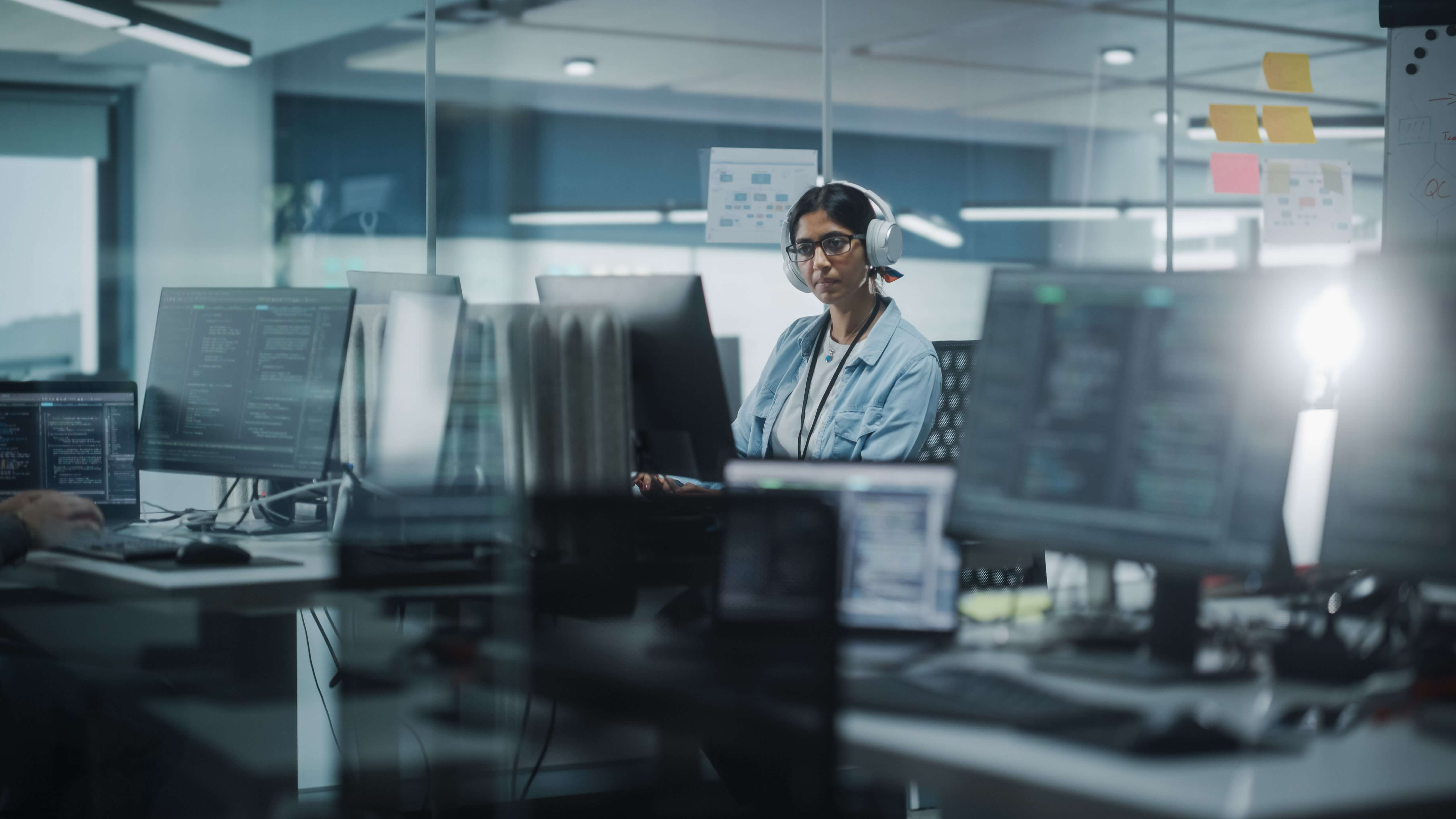 A woman working at her computer in a laboratory office, seen through glass partitions, with multiple computer monitors and office equipment in the foreground.