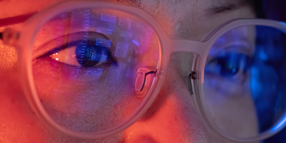 Close up of a woman's eyes, wearing glasses. Reflection of screen can be seen in her glasses in blue. 